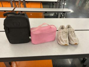 A black backpack, a pink lunchbox, and a pair of white sneakers are placed on a table in a cafeteria.