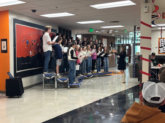 A choir performs on a stage in a school hallway, led by a conductor, with students and spectators present.