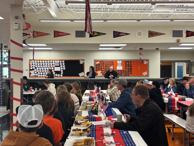 A school cafeteria filled with people enjoying a meal, decorated with flags and banners for a festive event.