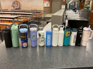 A row of ten assorted water bottles on a cafeteria table, featuring various colors and designs.