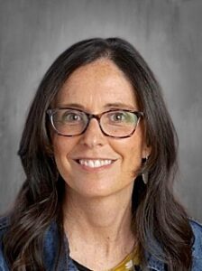 A smiling woman with long dark hair and glasses, wearing a denim jacket over a yellow top, against a gray background.
