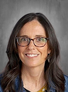A smiling woman with long dark hair and glasses, wearing a denim jacket over a yellow top, against a gray background.