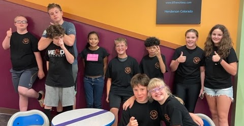 A group of ten children in matching black t-shirts pose together in a colorful indoor setting, smiling and giving thumbs up.