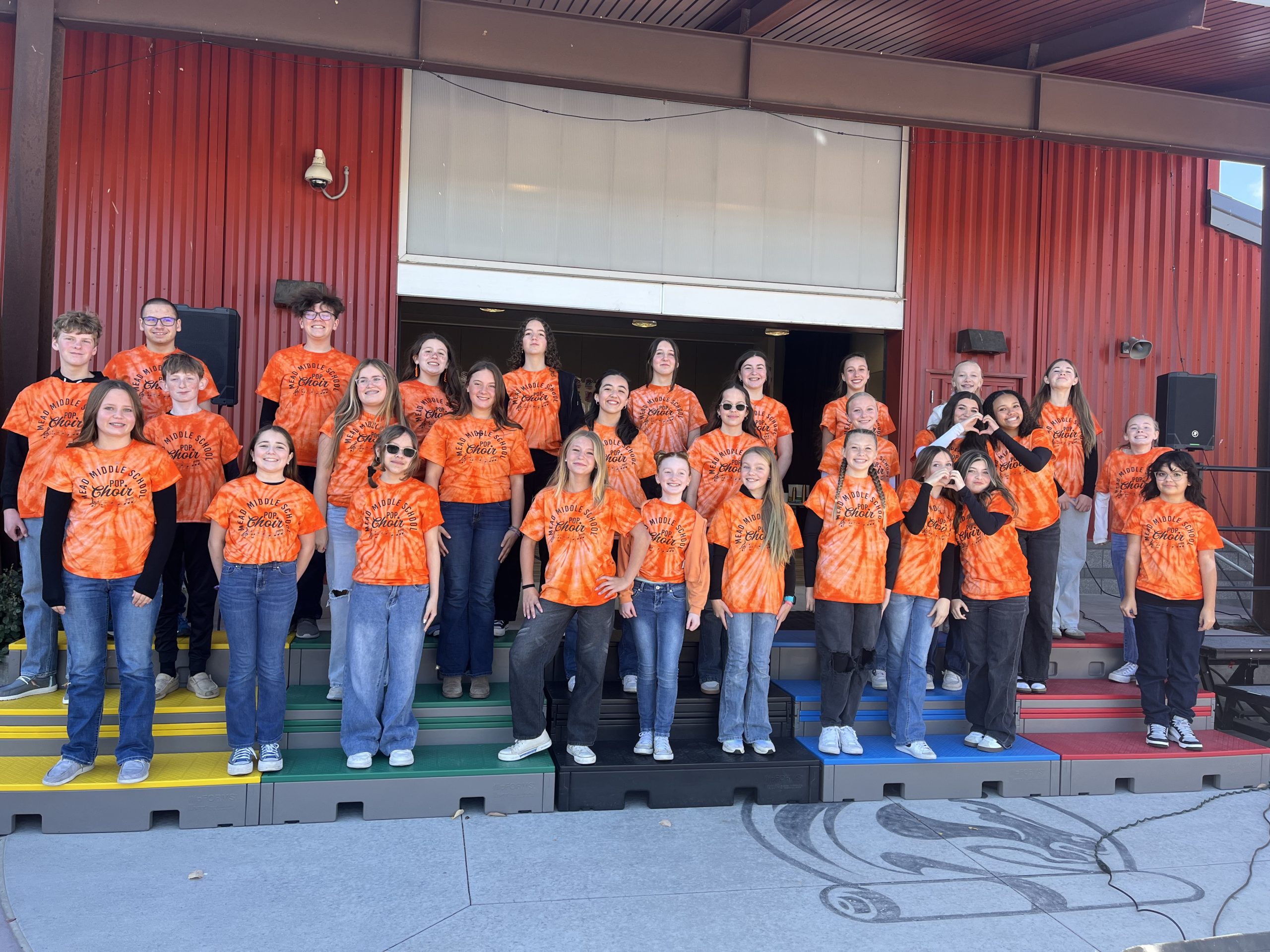 Un grupo de estudiantes con camisetas naranjas teñidas al estilo «tie-dye» posa en unas escaleras de colores frente a un edificio rojo, sonriendo y divirtiéndose.
