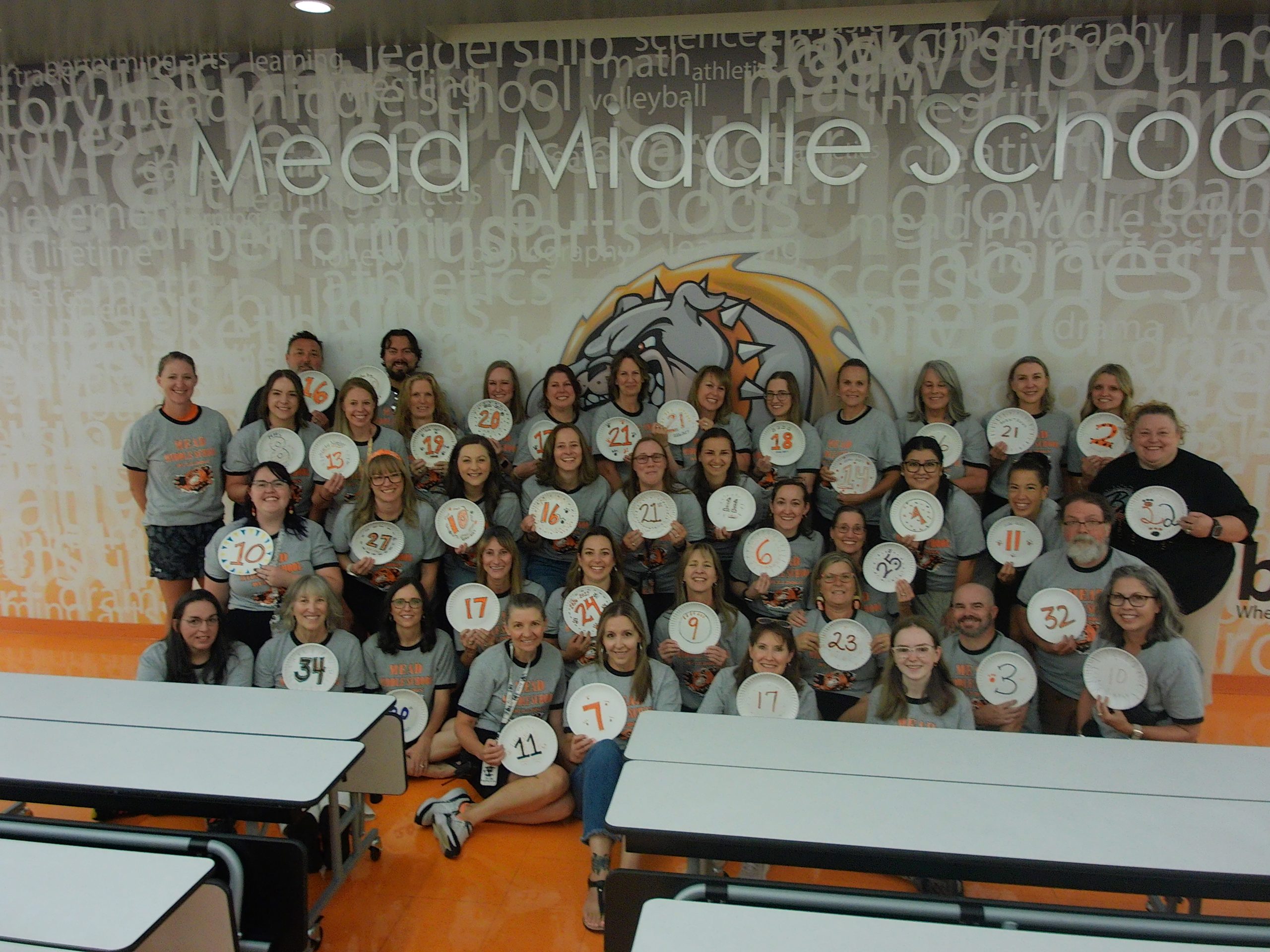 Un grupo de profesores y personal de la escuela « Mead Middle » posa juntos, sosteniendo placas con números, en el comedor de la escuela.